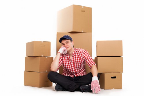 A young man dressed in a red and white checkered shirt, wearing a navy cap and pink packing gloves, is sitting cross-legged on the floor in front of a collection of six cardboard moving boxes of varying sizes, arranged in a semi-circle around him. The boxes are plain brown with no visible labels or markings, and some are stacked, with a large box situated directly behind the man. The background is a plain white, emphasizing the boxes and the man's relaxed, friendly expression as he rests his head on his left hand. The scene appears to be indoors, likely in a residential setting typical of a house or flat in Camden, with ample natural or studio lighting highlighting the cardboard textures and the man's clothing. The image visually represents a house removal or packing process, fitting within the context of Camden Man with Van’s removals services, illustrating readiness for a move in the local area.
