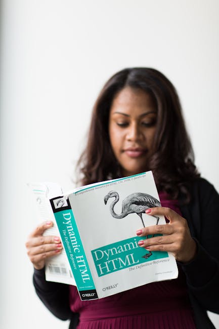 A woman with dark hair, dressed in a burgundy top and black blazer, stands indoors holding an open technical book titled 'Dynamic HTML' by O'Reilly with a teal cover and an illustration of a pink flam