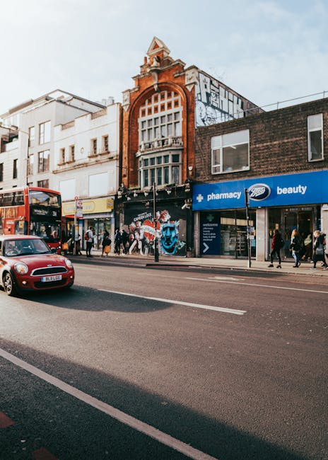 A street scene on Kentish Town Road featuring a brick building with large arched and rectangular windows, some with window blinds, and a clock mounted on a black pole in front. To the right, there is 