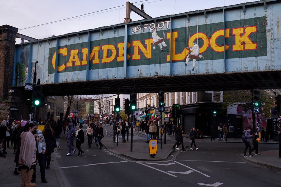 A busy street scene in Camden Town, NW1, showing pedestrians walking along the pavement and crossing the road, with a variety of red-brick and ornate Victorian-style buildings lining both sides of the